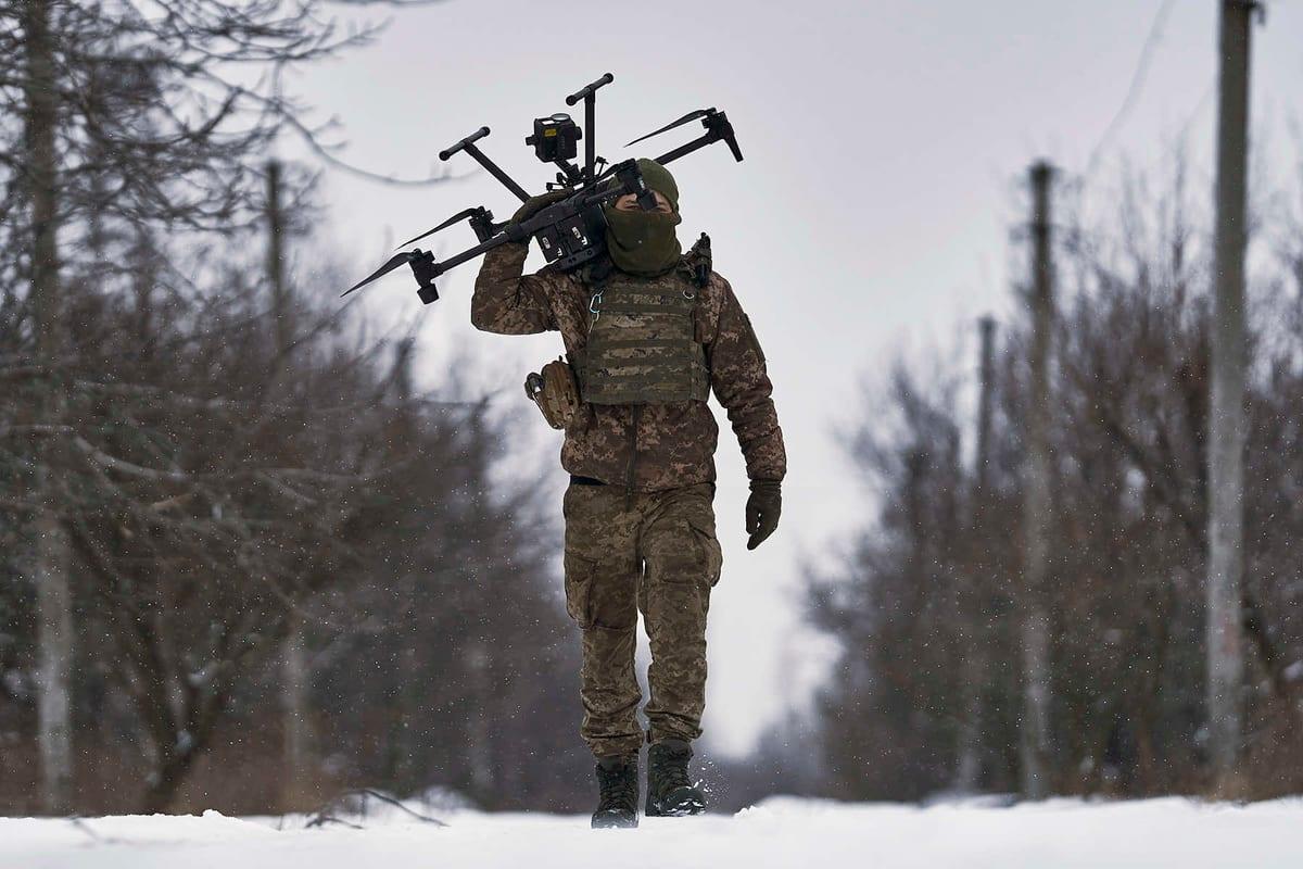 A soldier in winter camouflage carrying a large military drone along a snowy road