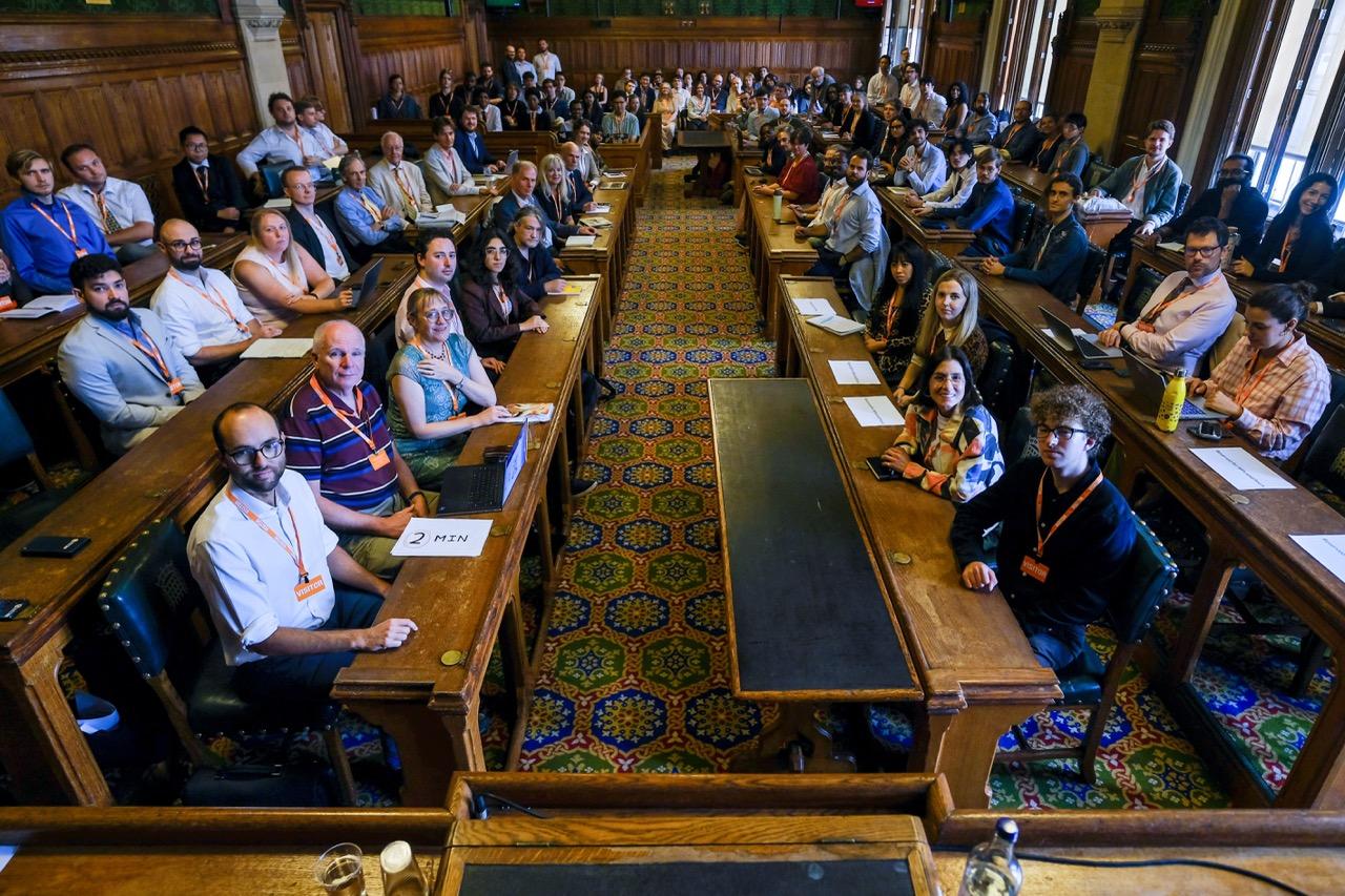 A packed committee room in the Houses of Parliament, with attendees at wooden benches on both sides of the central aisle turned to face the camera