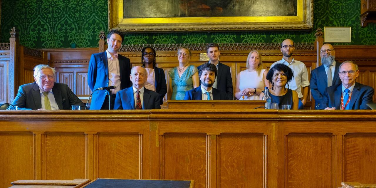 The panel seated at a long wooden bench in a wood-panelled committee room in the Houses of Parliament, with PauseAI UK organisers standing behind them